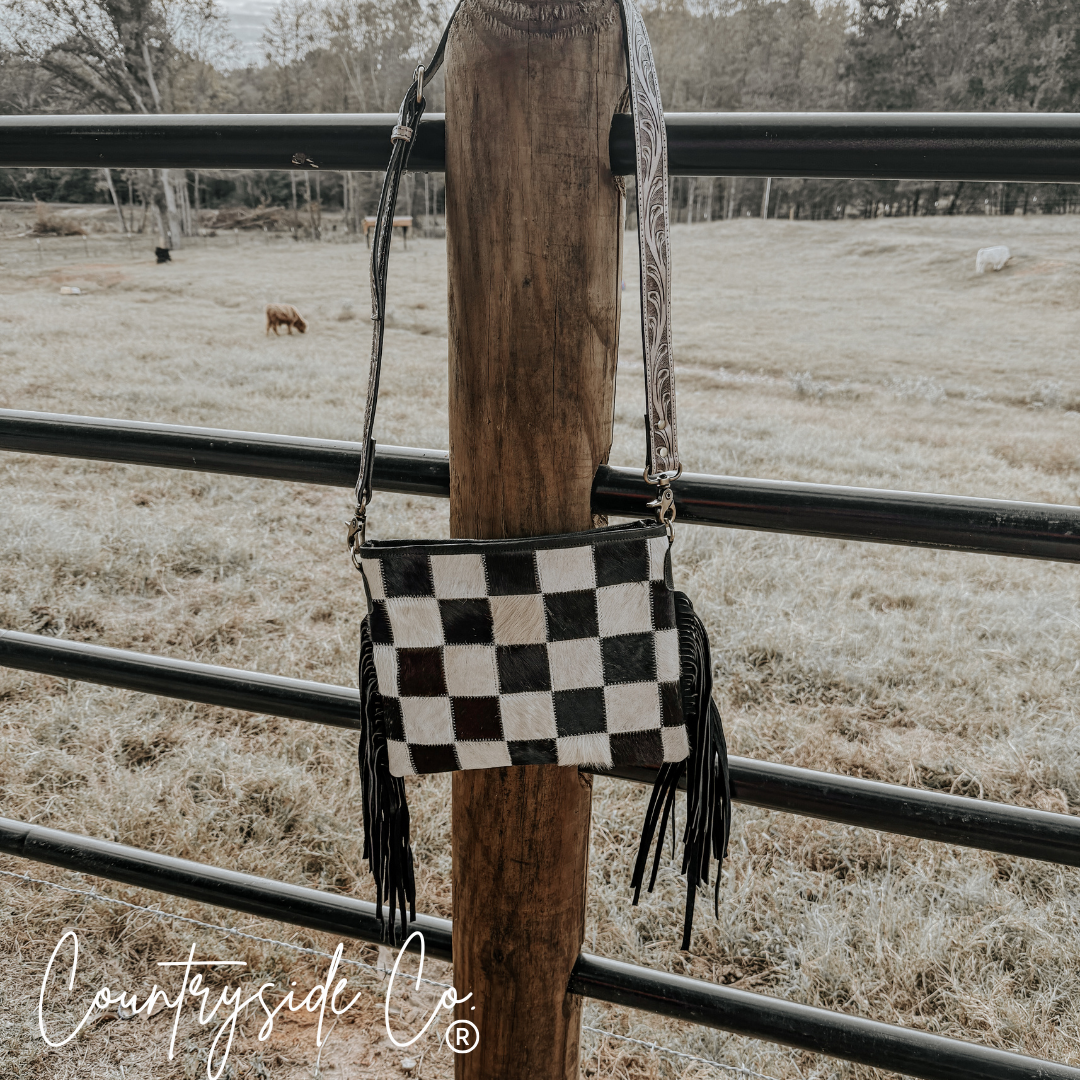 Rodeo Square Cowhide Checkered Purse hanging on a post in a rustic outdoor setting with a checkered design and fringes.