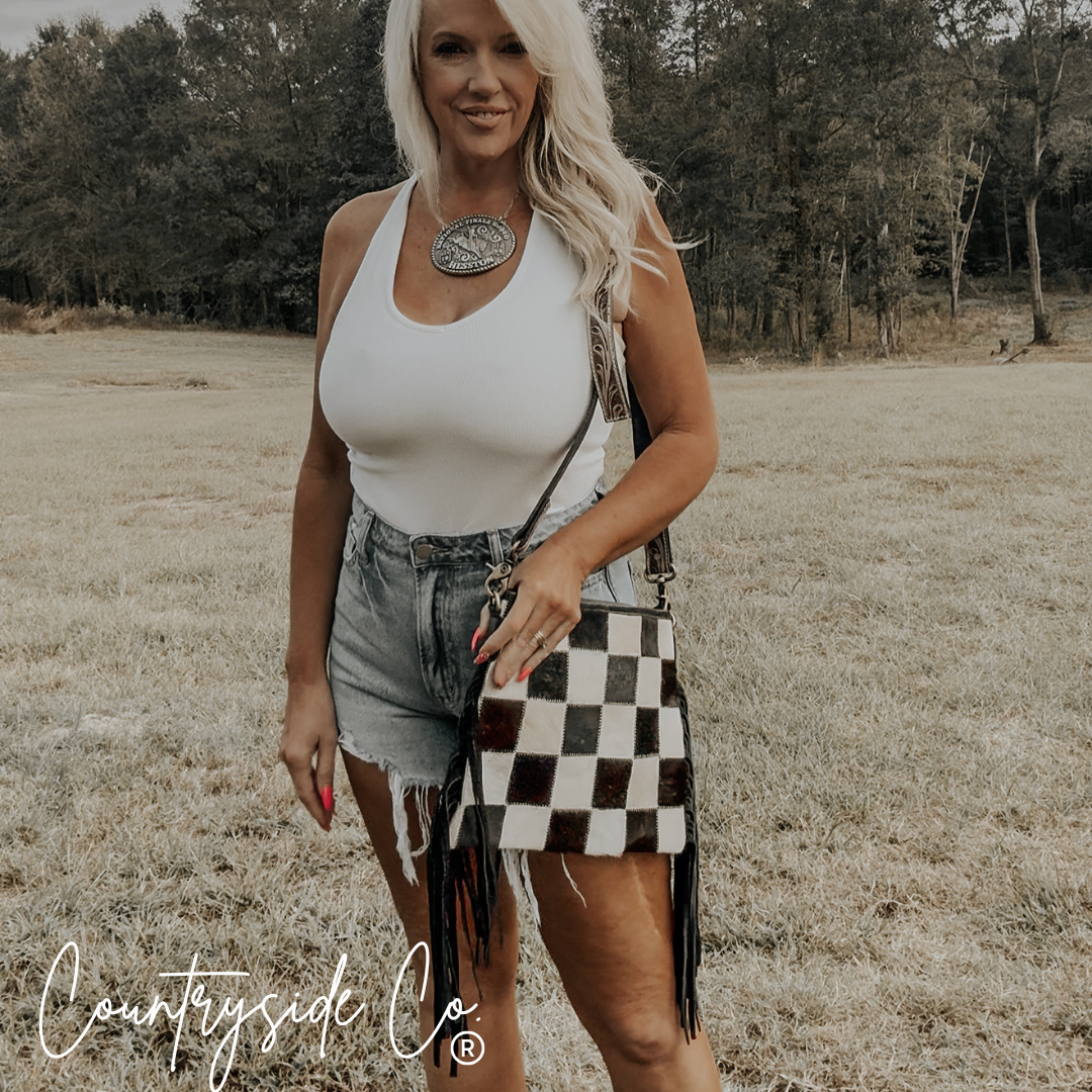 Woman holding a Rodeo Square Cowhide Checkered Purse in a country setting, showcasing western style and charm.