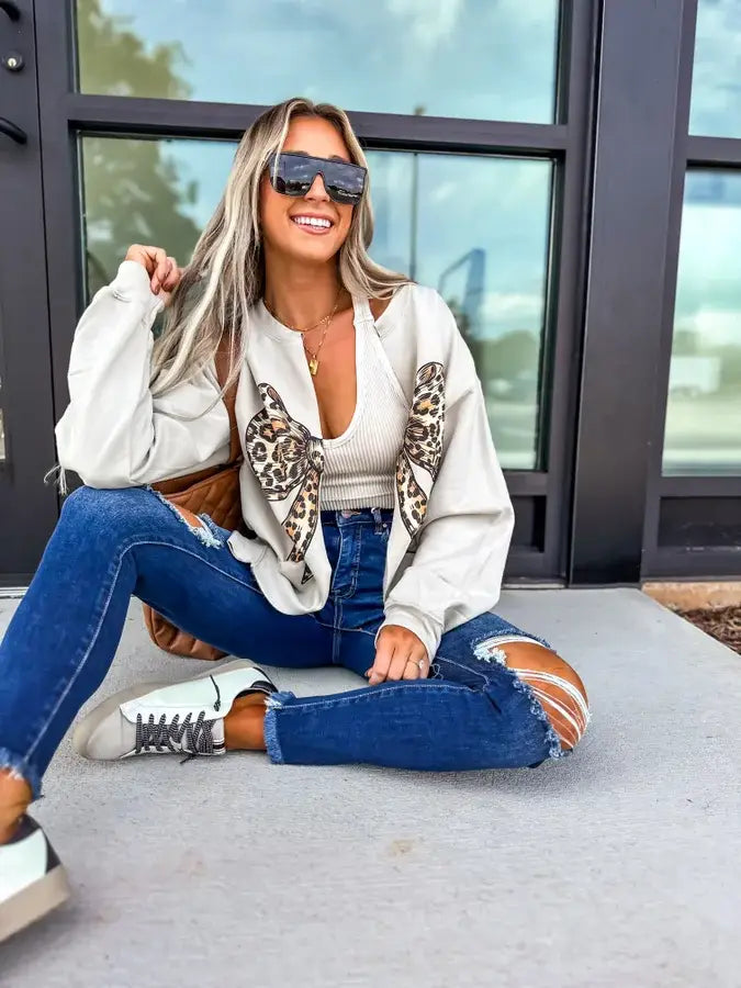 Leopard Bow Sand Graphic Cardigan worn by a smiling woman with sunglasses, paired with ripped jeans and casual shoes.