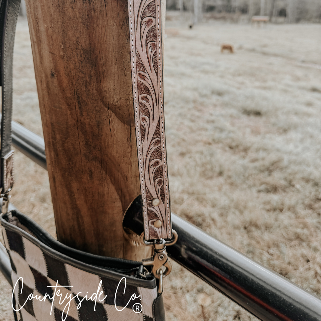 Detail of the tooled leather strap on Rodeo Square Cowhide Checkered Purse hanging on a post outdoors.