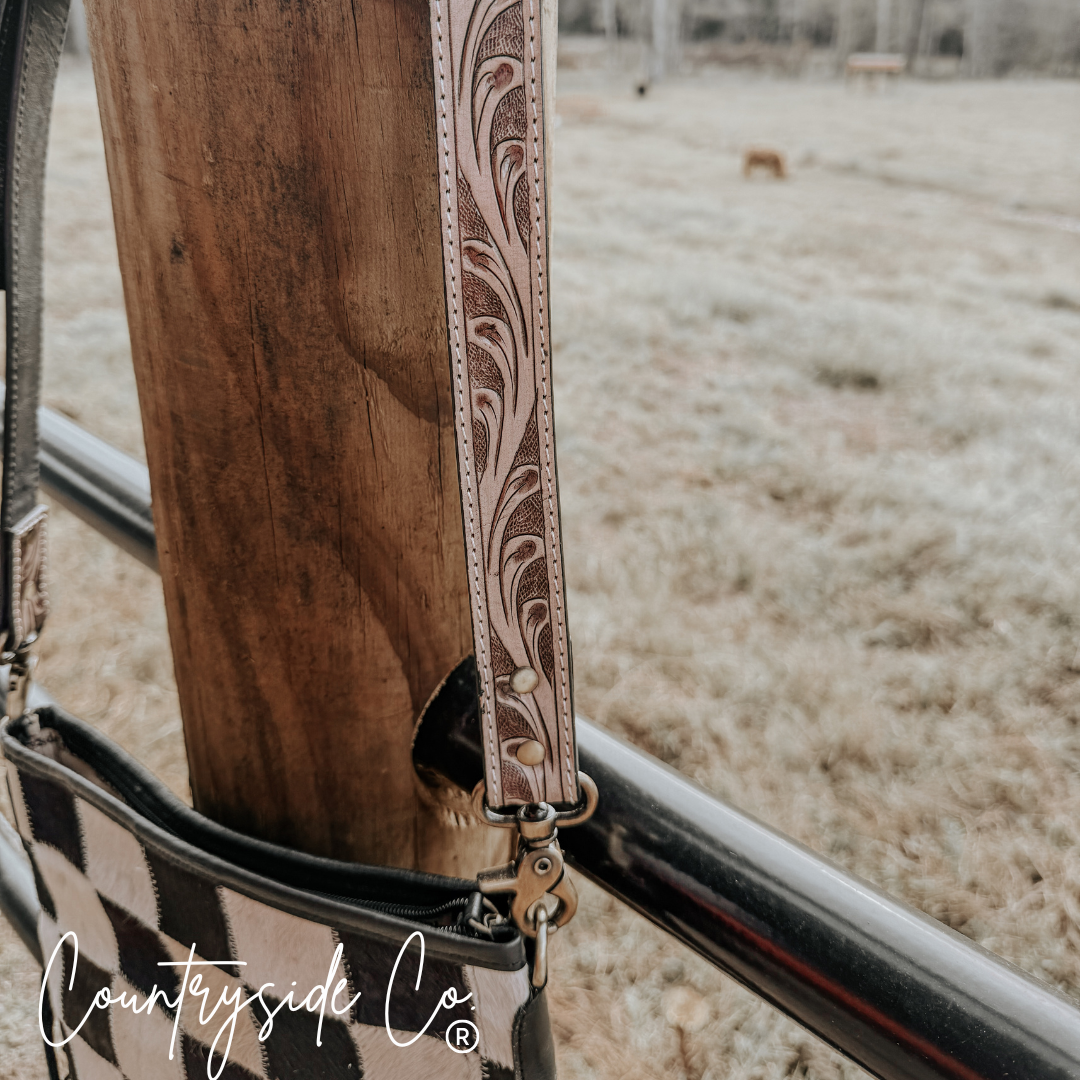 Detail of the tooled leather strap on Rodeo Square Cowhide Checkered Purse hanging on a post outdoors.