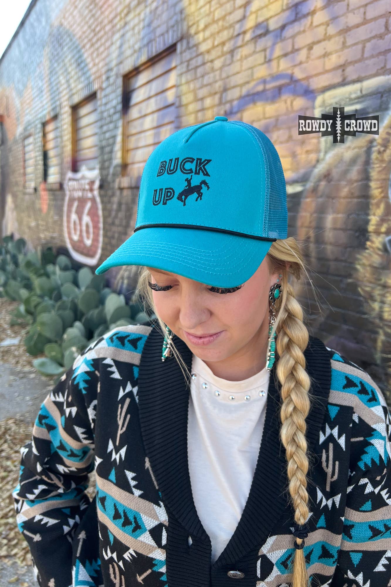 Woman wearing a teal Buck Up Cap with embroidered design standing in front of a colorful mural.